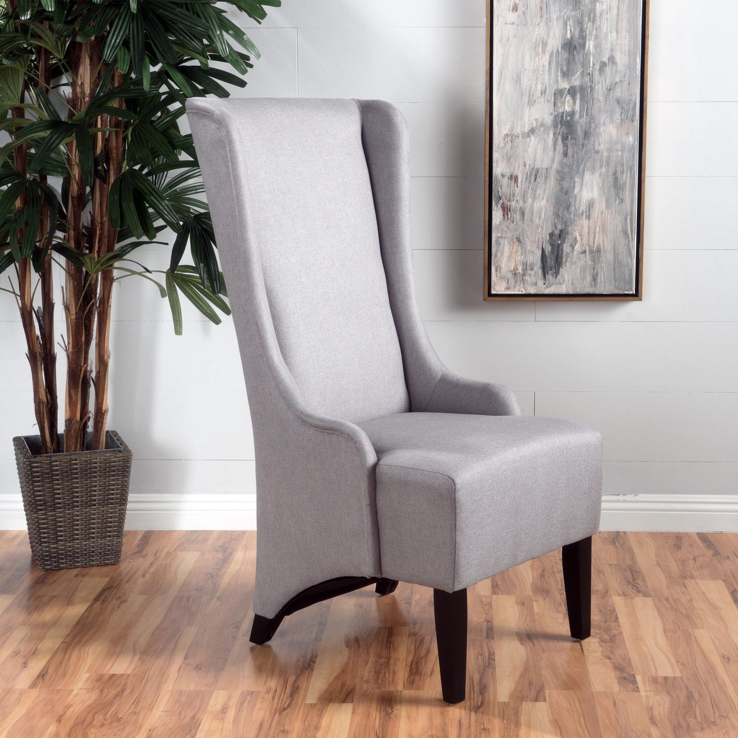 An Alight Upholstered Dining Chair with a high back and birch wood legs sits on a wood floor near a potted plant and a framed abstract painting on a white wall.