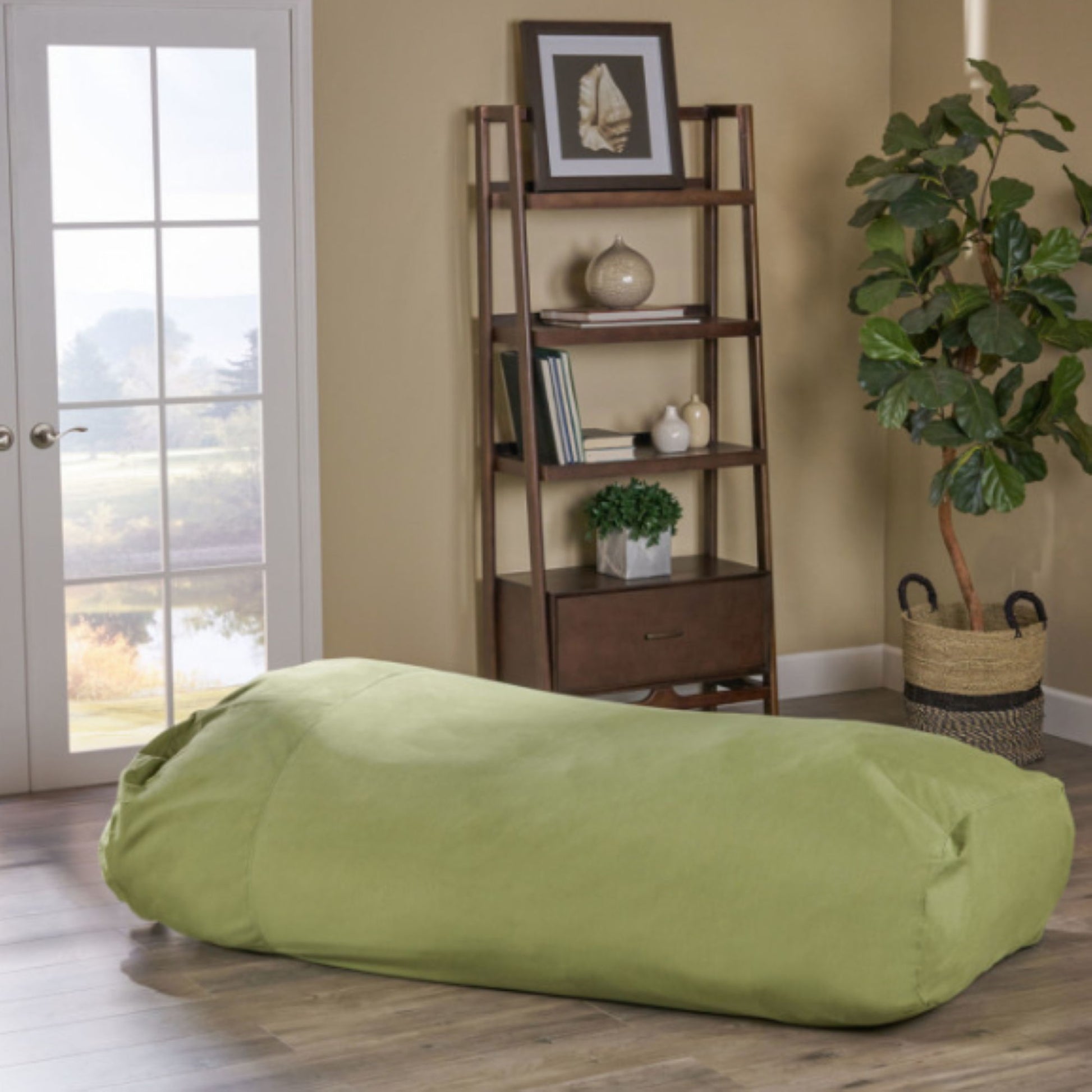 The Kasper - Water Resistant Lounger Bean Bag in light green rests on a wooden floor beside a tall potted plant, a wooden shelf with books and decor, and a large window filling the cozy room with natural light.