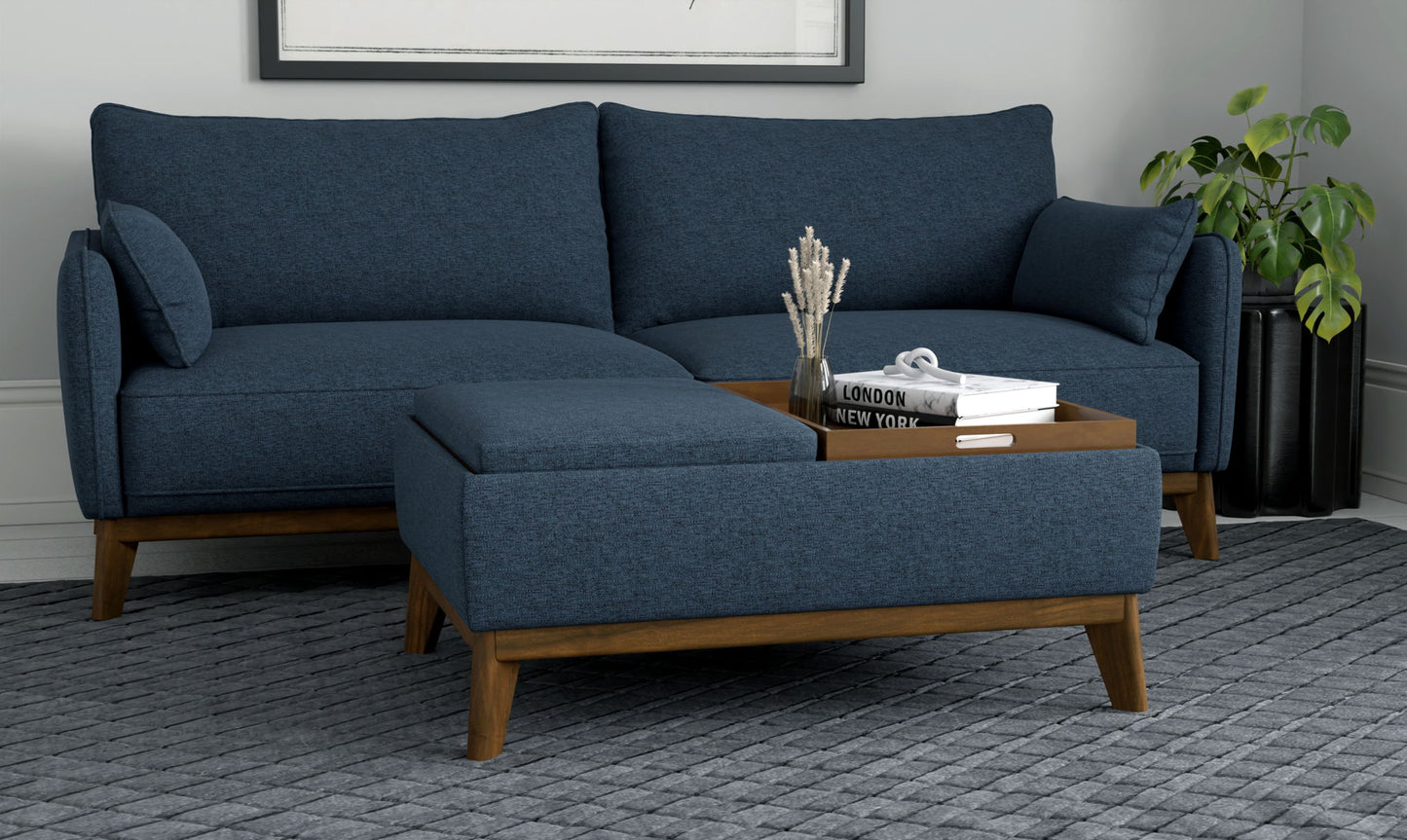A modern living room featuring a blue sectional sofa, wooden legs, the Varia Reversible Tray Ottoman in blue with decor items, a gray textured rug, and a green plant in the corner.