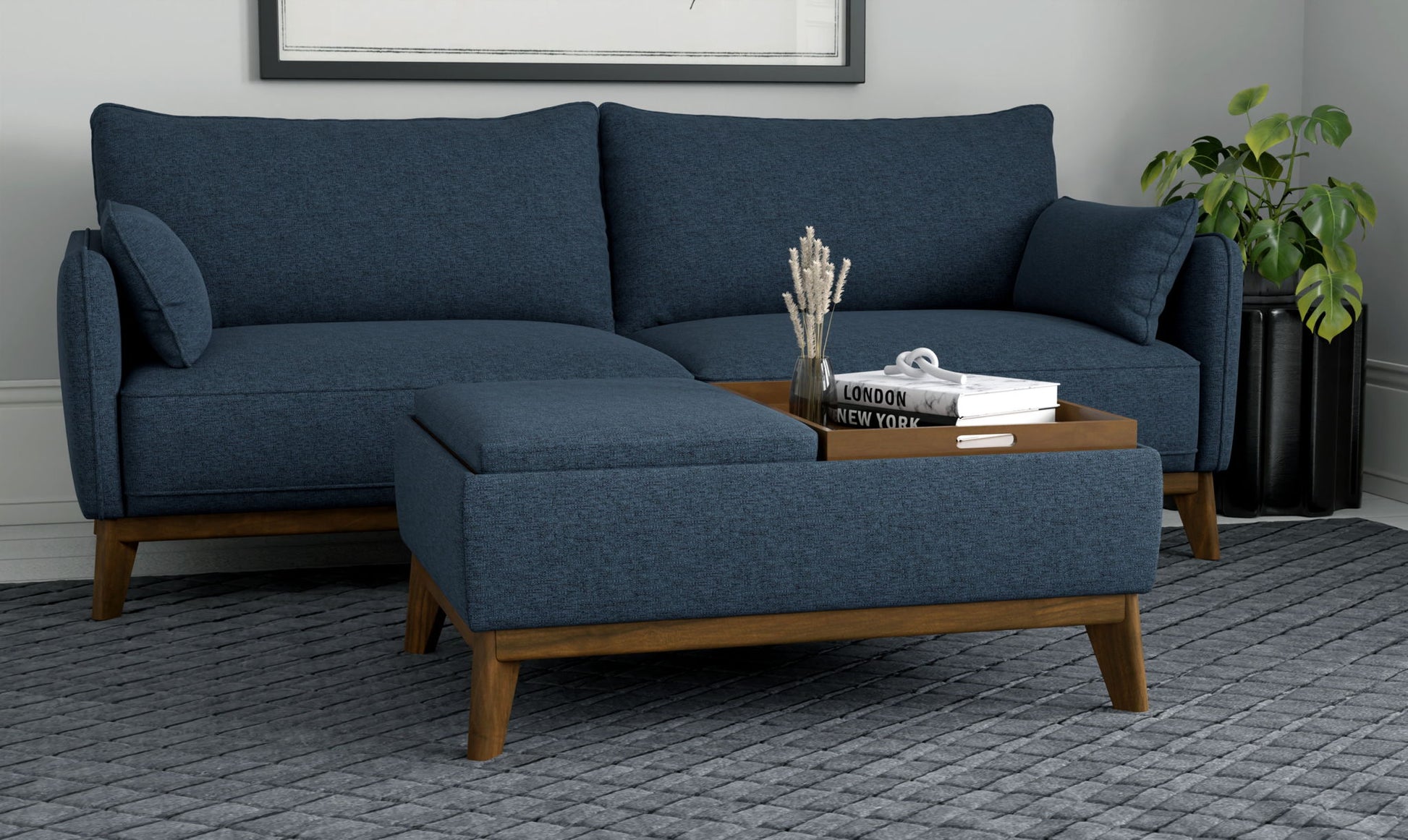 A modern living room featuring a blue sectional sofa, wooden legs, the Varia Reversible Tray Ottoman in blue with decor items, a gray textured rug, and a green plant in the corner.
