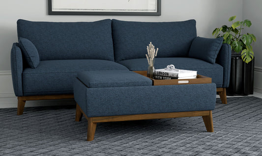 A modern living room featuring a blue sectional sofa, wooden legs, the Varia Reversible Tray Ottoman in blue with decor items, a gray textured rug, and a green plant in the corner.