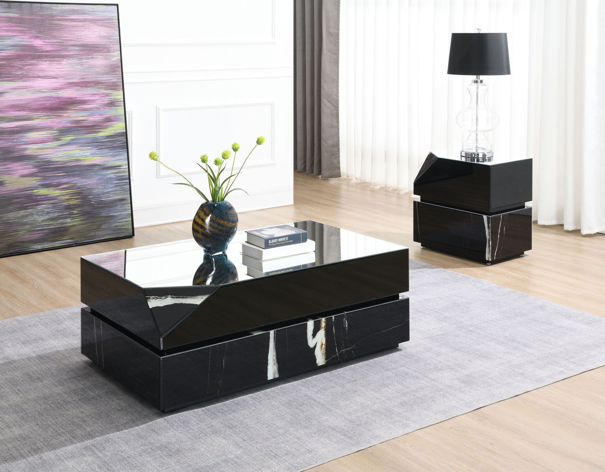 A modern living room featuring a glossy black coffee table, stacked books, a vase with green stems, the Drisana Mirrored End Table in black with a lamp, a large abstract painting, light wood flooring, and a gray rug.