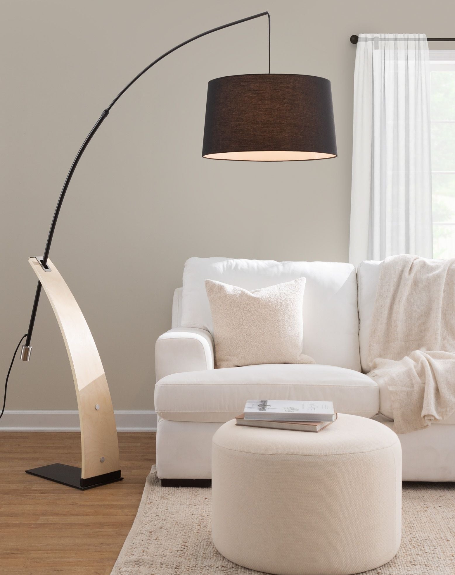 A modern living room with a white sofa, beige cushions, a Large Round Pouf - Knitted Beige, light beige throw blanket, and a tall black and wood arc floor lamp, set against light walls and a window with white sheer curtains.