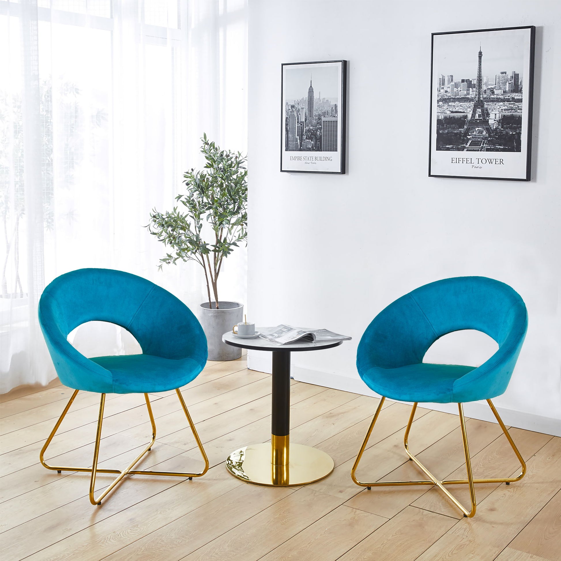 Two Modern Accent Velvet Lounge Chairs in blue with gold legs flank a small round table with a black and gold base, set by a large window. A potted plant and framed art decorate the white wall behind.