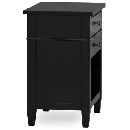 The Carlton Bedside Table is a handcrafted black wooden nightstand featuring one drawer with bronze knobs, an open lower shelf, simple modern design with straight lines, and four sturdy legs.