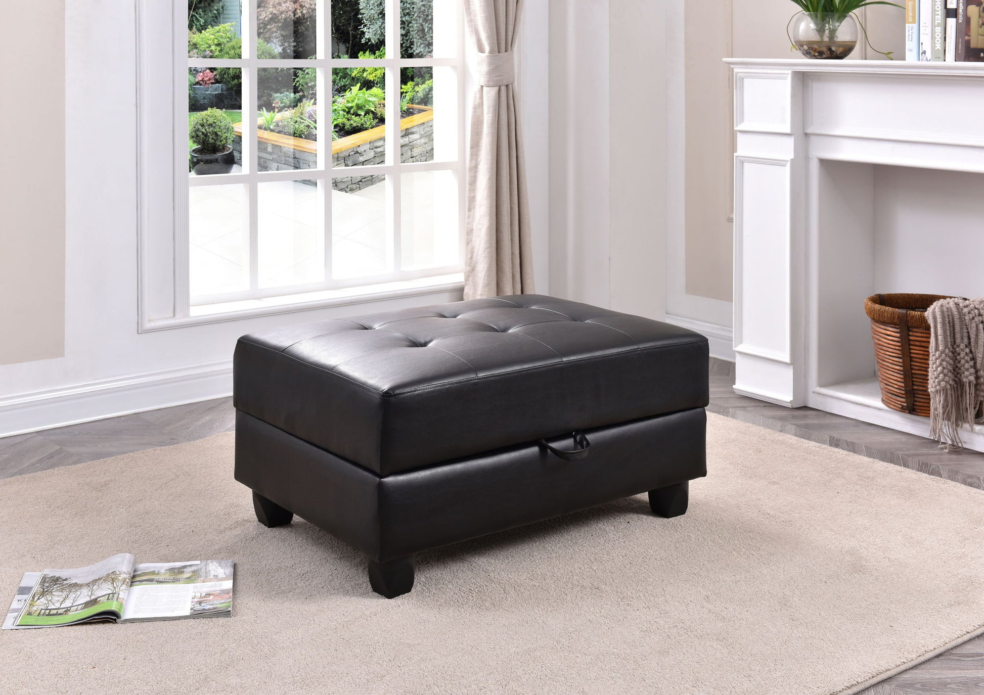 The Stylish Storage Contemporary Ottoman in black leather, featuring tufted details, sits on a beige rug in a bright living room with large windows, a white fireplace, indoor plants, and an open magazine on the floor.