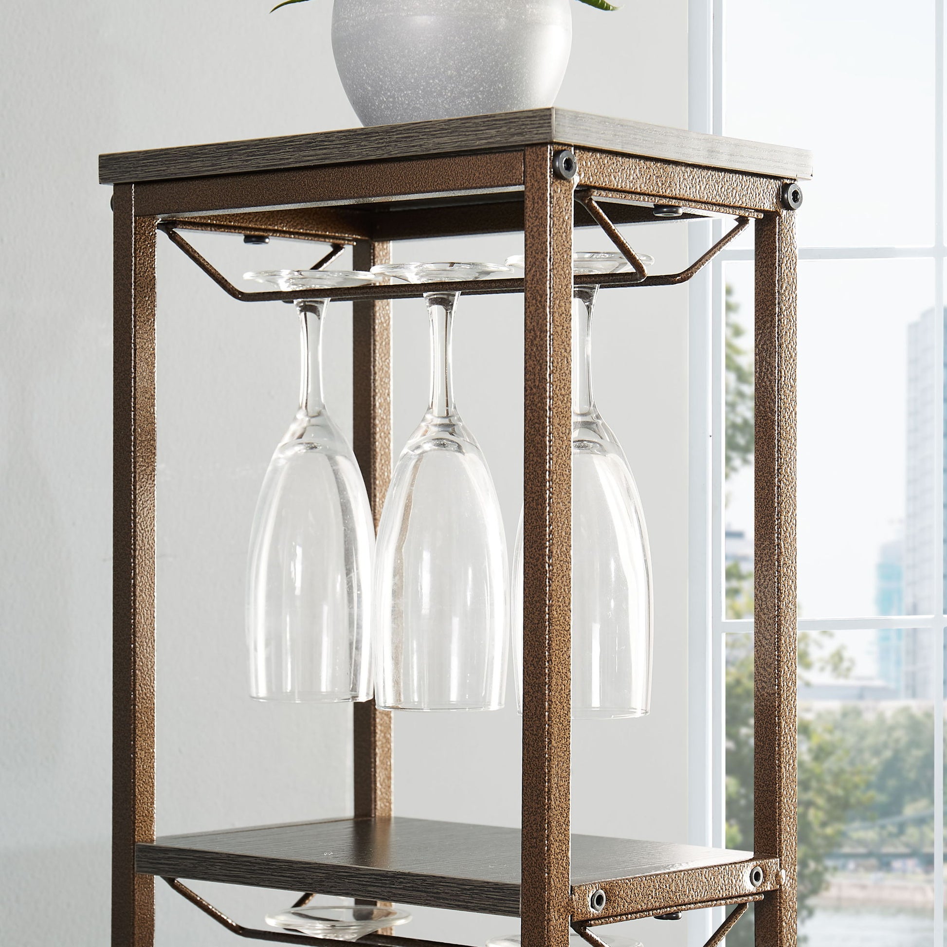 A close-up of the 11 Bottle Wine Bakers Rack, featuring a hanging wine glass holder with four upside-down glasses, wood and metal shelves, a white planter on top, and a window in the background.