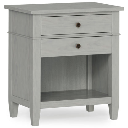 The Carlton Bedside Table is a handcrafted, light gray wooden nightstand with two drawers featuring round metal knobs, an open lower shelf, and simple tapered legs for a modern, minimalist look.