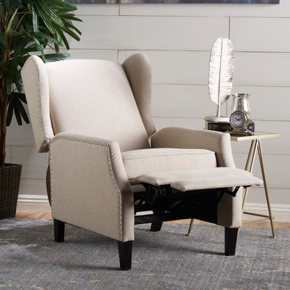 The Westeros Wing Back Fabric Upholstered Recliner, in beige with nailhead trim, is partially reclined on a gray rug beside a side table holding a lamp, clock, and plant, all set against a white shiplap wall.
