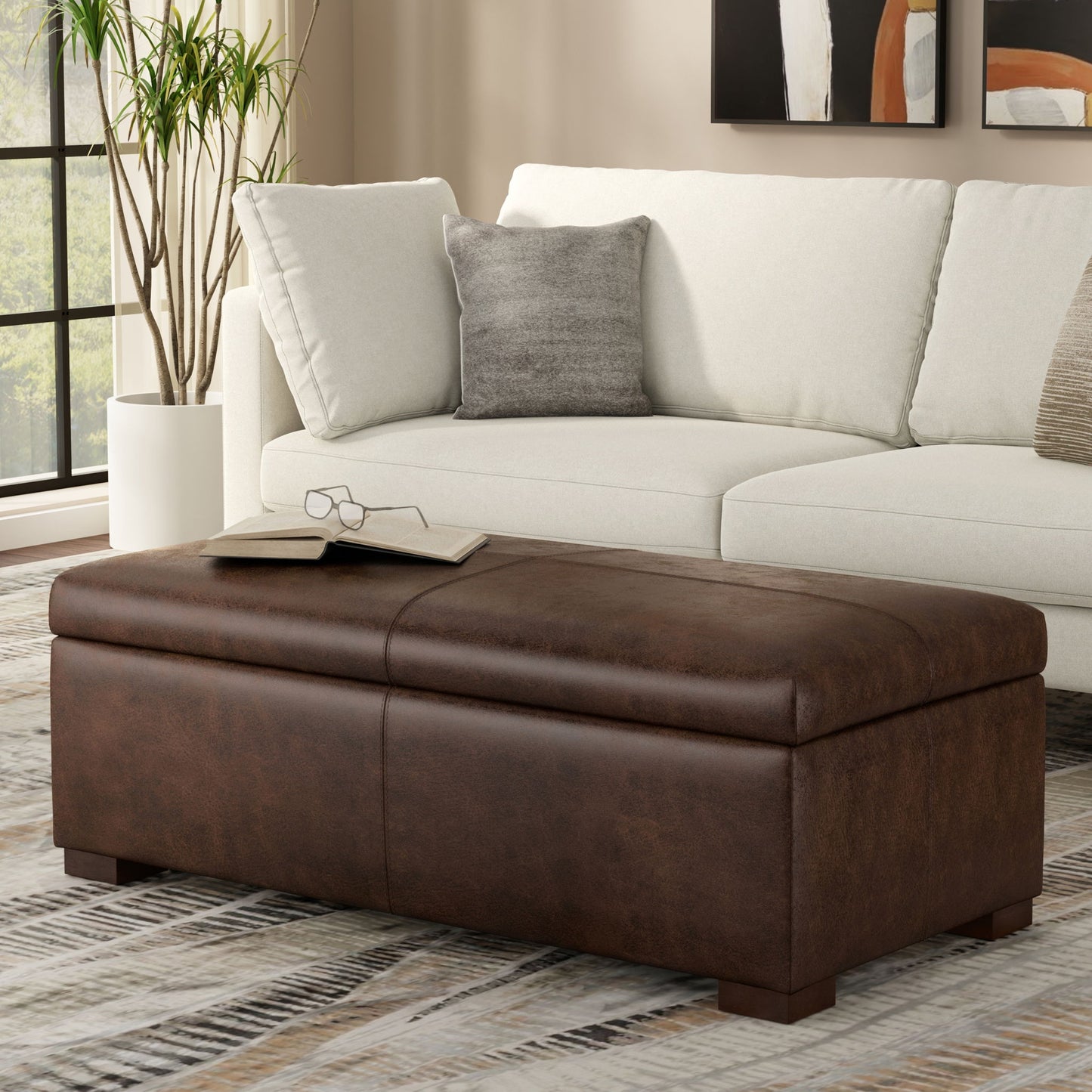 The Gabbie Coffee Table Upholstered Storage Ottoman with glasses on top sits before a light-colored sofa with cushions in a modern living room featuring large windows and a patterned rug.