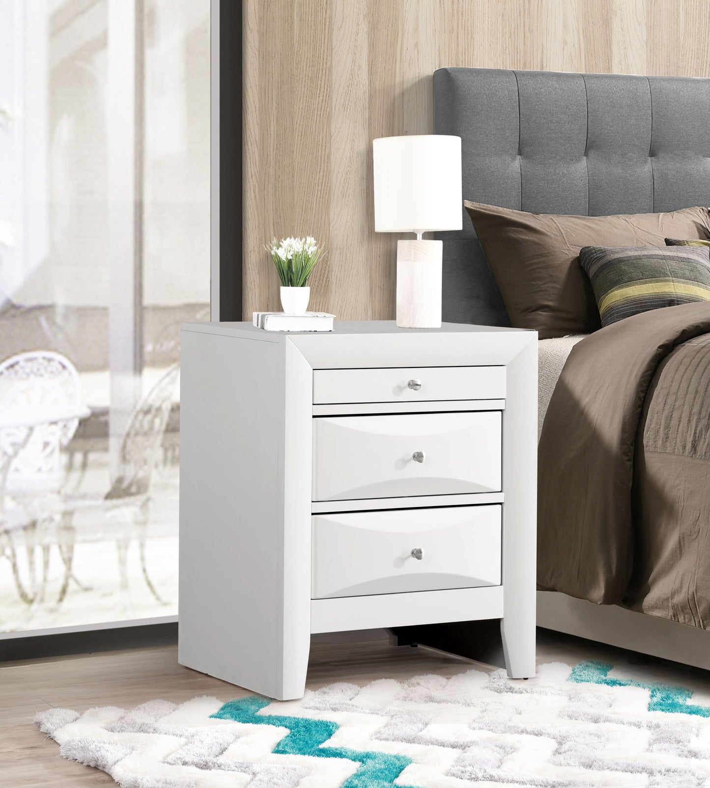 The Marilla Nightstand, a modern white piece with three drawers, is placed beside a bed dressed in gray and brown bedding. The tabletop holds a white lamp and small potted plant, against a wood-paneled wall and patterned rug.