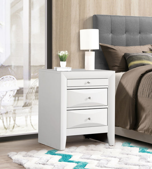 The Marilla Nightstand, a modern white piece with three drawers, is placed beside a bed dressed in gray and brown bedding. The tabletop holds a white lamp and small potted plant, against a wood-paneled wall and patterned rug.