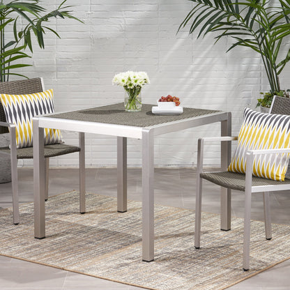 Two modern patio chairs with yellow and gray pillows are placed at the Outdoor Square Dining Table With Aluminum Frame, which holds a vase of white flowers and grapes, all set on a rug with green plants in the background.