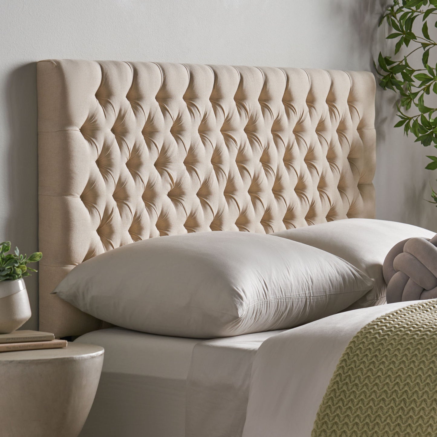 The Annecy - Contemporary Upholstered Headboard in beige features a luxurious, quilted design behind a neatly made bed with light bedding, pillows, and a textured green blanket, beside a round table and leafy plant.