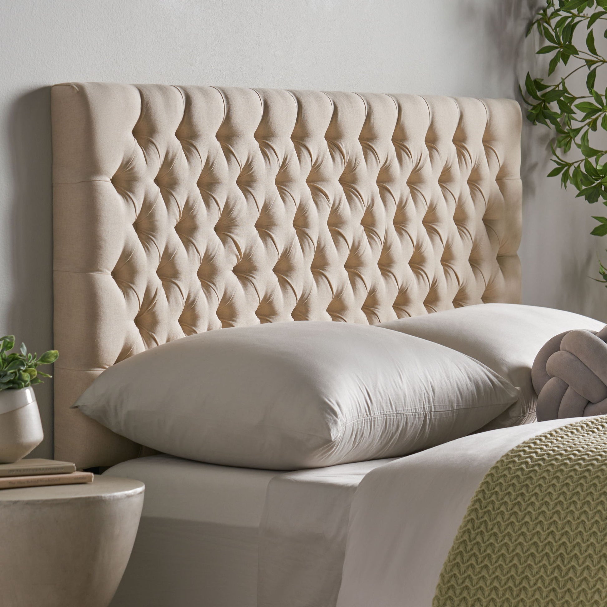 The Annecy - Contemporary Upholstered Headboard in beige features a luxurious, quilted design behind a neatly made bed with light bedding, pillows, and a textured green blanket, beside a round table and leafy plant.