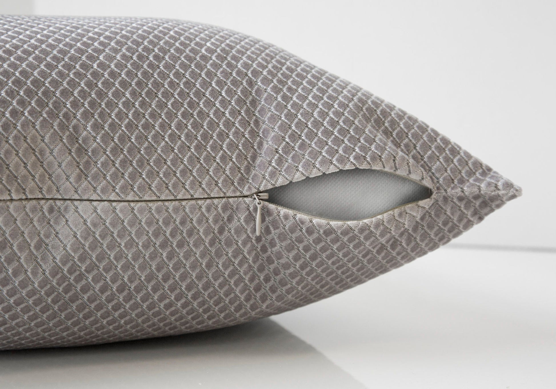 A modern, decorative square pillow with a gray textured cover and visible zipper, partially open to show the white hypoallergenic insert. Pillow is resting on a smooth, white surface.