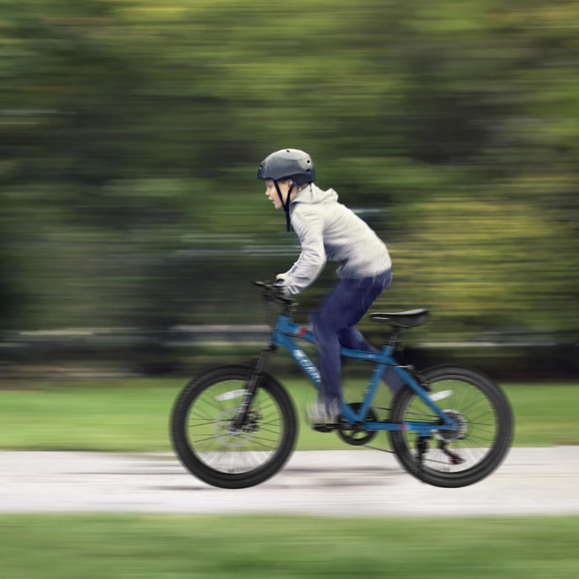 A person in a helmet and grey hoodie rides the S20101 20" Kids' Bike, a blue 7-speed mountain bike for ages 8-12 with front suspension and disc brake, quickly along a path with a blurred green background indicating motion.