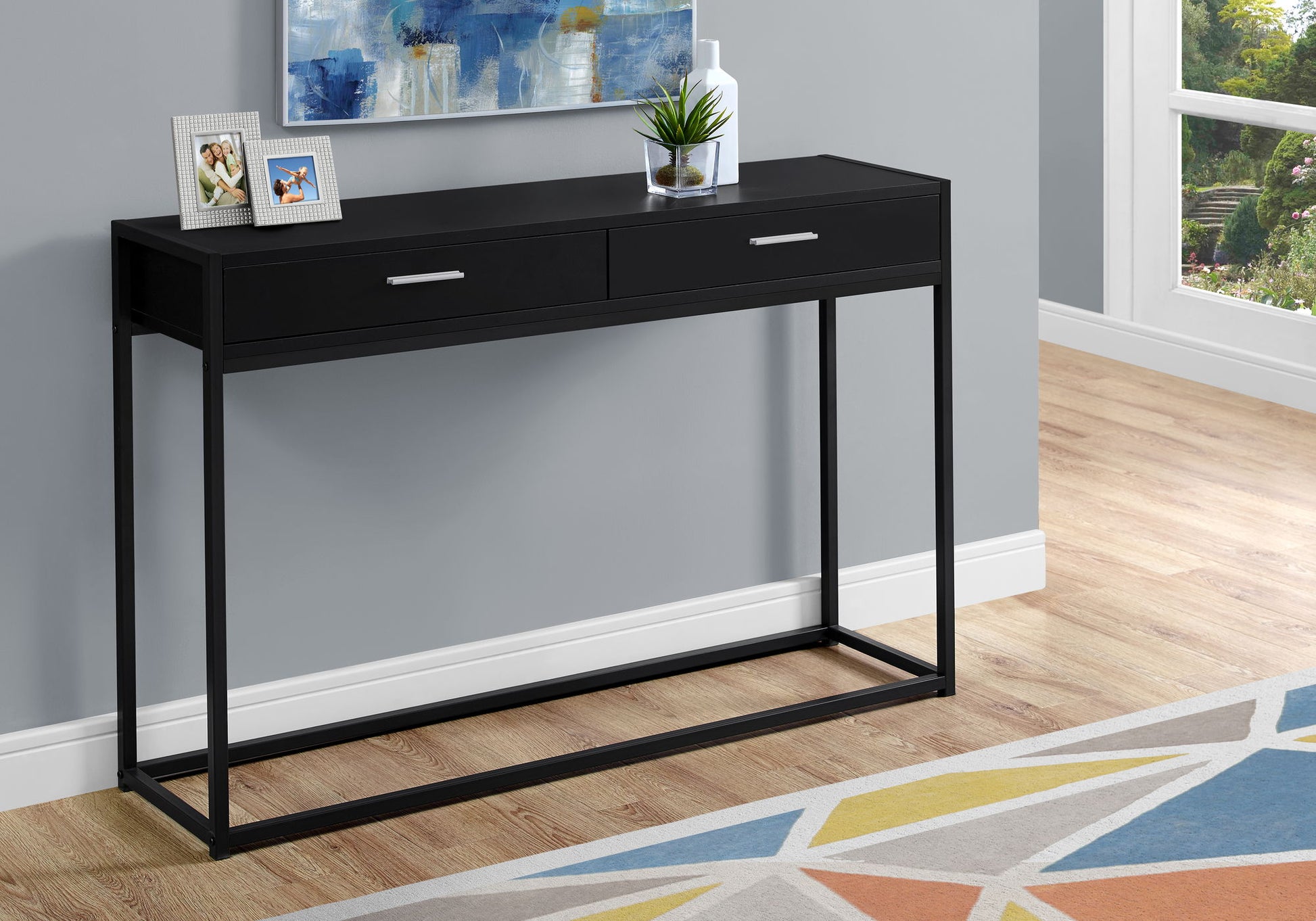 The Accent Console Table For Entryway with storage drawers sits against a gray wall, topped with a framed photo, potted plant, and vase. A colorful rug and a window overlooking the garden complete the contemporary scene.