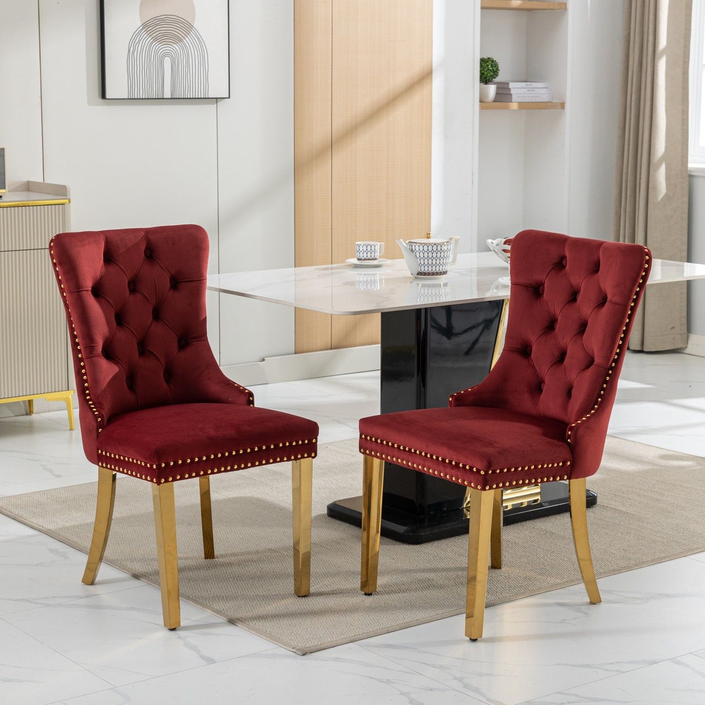 The Nikki set of two modern velvet dining chairs with tufted backs, nailhead trim, and gold-plated legs complement a contemporary dining room featuring a marble table, light rug, shelves, and framed wall art.