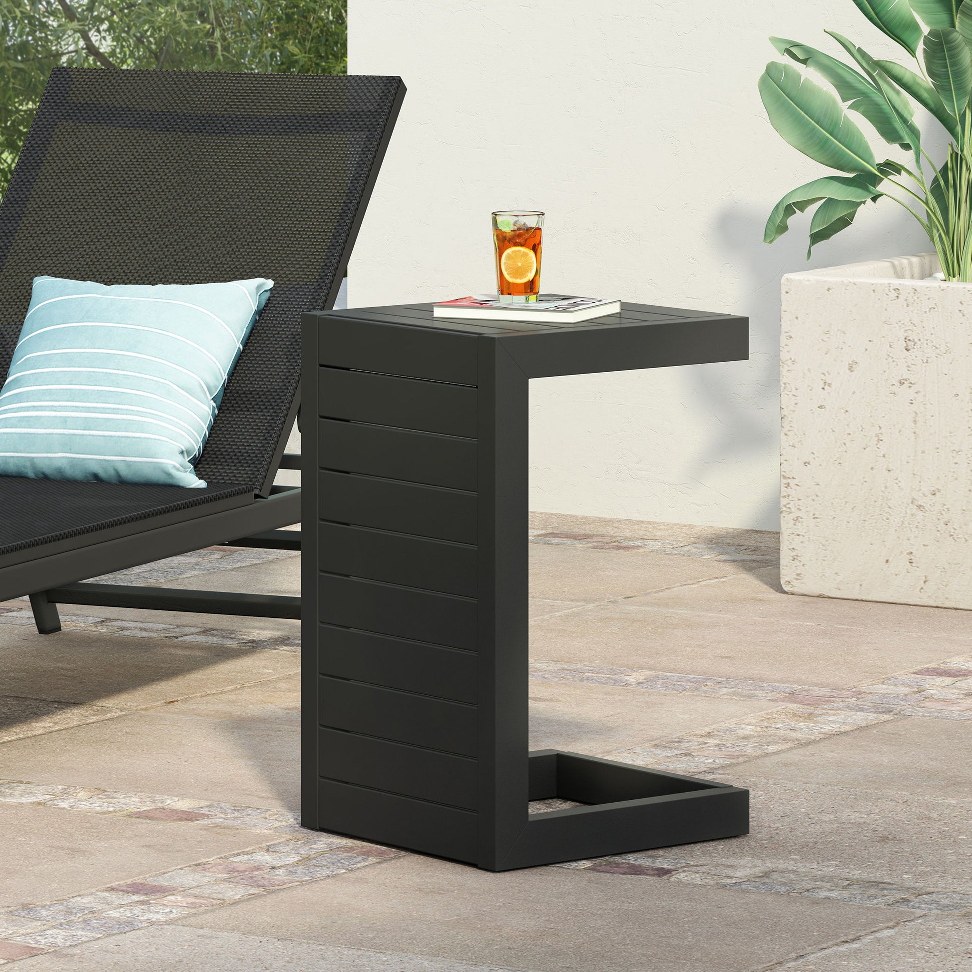 An Aluminum Outdoor C-Shaped Side Table for Patio in black stands beside a lounge chair with a striped pillow, holding a glass of iced tea with citrus slices. Nearby, a large planter with green leaves decorates the stone patio.