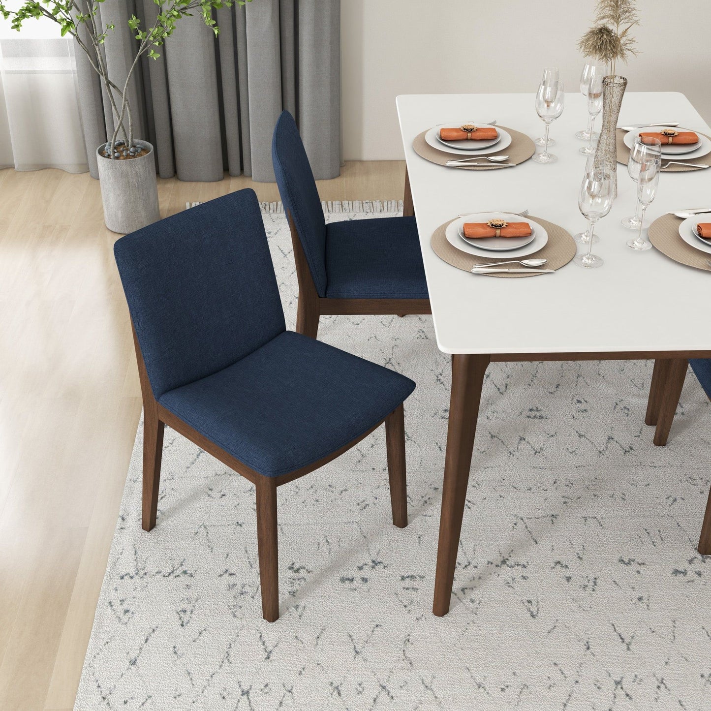 A modern dining room with a white rectangular table set for four, featuring Laura Mid-Century Modern Solid Wood Dining Chairs in dark blue, a light patterned rug, and a potted plant by the window with gray curtains.