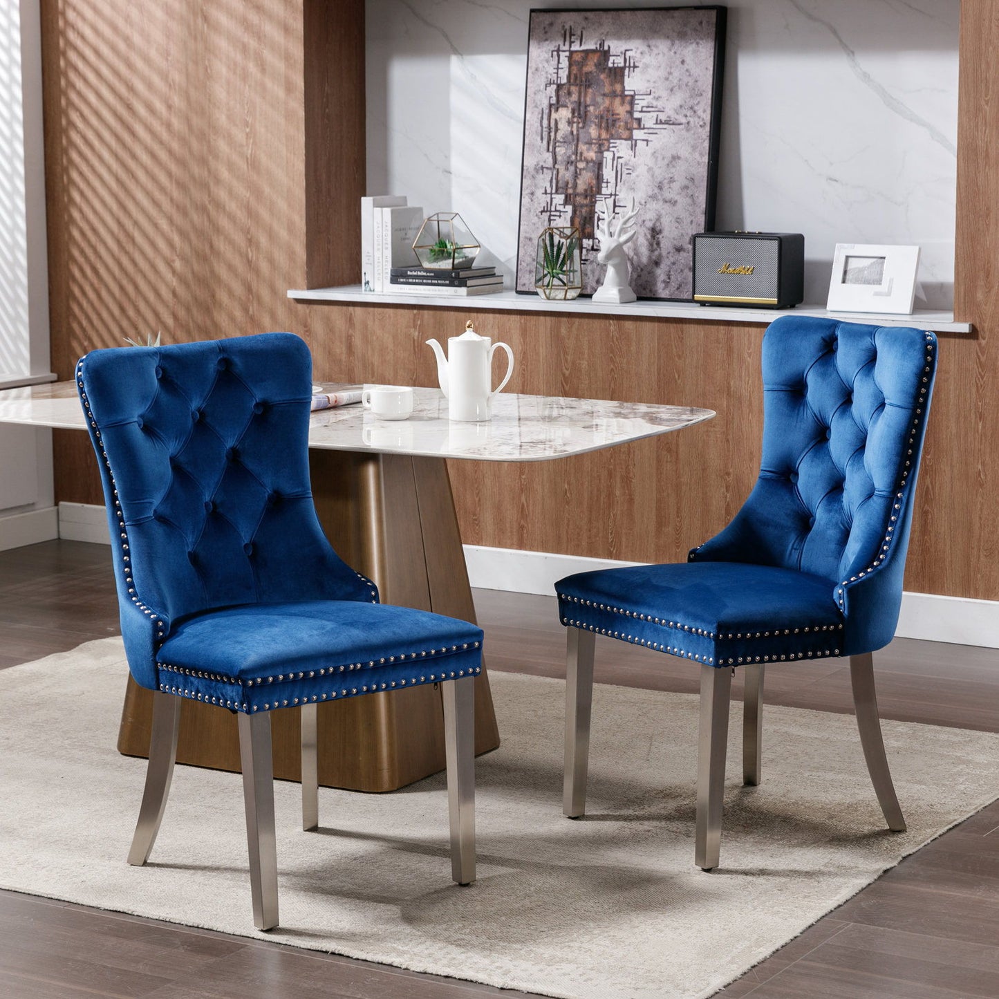A set of two Nikki modern velvet dining chairs with tufted backs, chrome stainless steel legs, and nailhead trim sit on a beige rug by a marble table in a stylish room with wood paneling, wall art, decor, and sunlight streaming in.