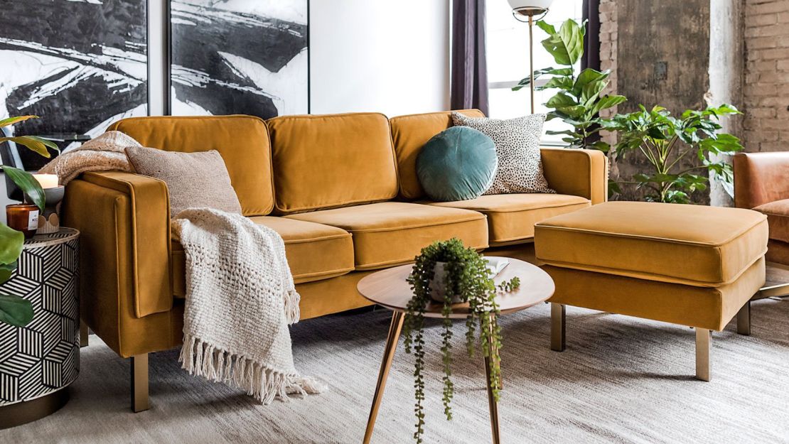 A modern living room featuring a mustard yellow couch with a matching ottoman. A knitted throw and decorative pillows adorn the couch, and a small wooden coffee table holds a leafy plant. The room is accented with a large abstract painting and numerous indoor plants.