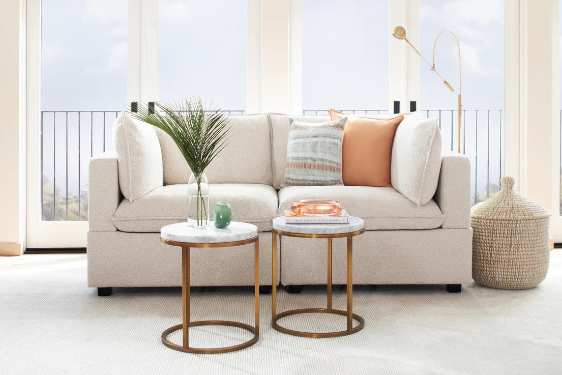 A cozy living room featuring a beige sofa adorned with decorative pillows. Two modern round side tables with marble tops and gold legs are placed in front of the sofa. A small plant, a candle, and a decorative object are on the tables. A floor lamp stands nearby.