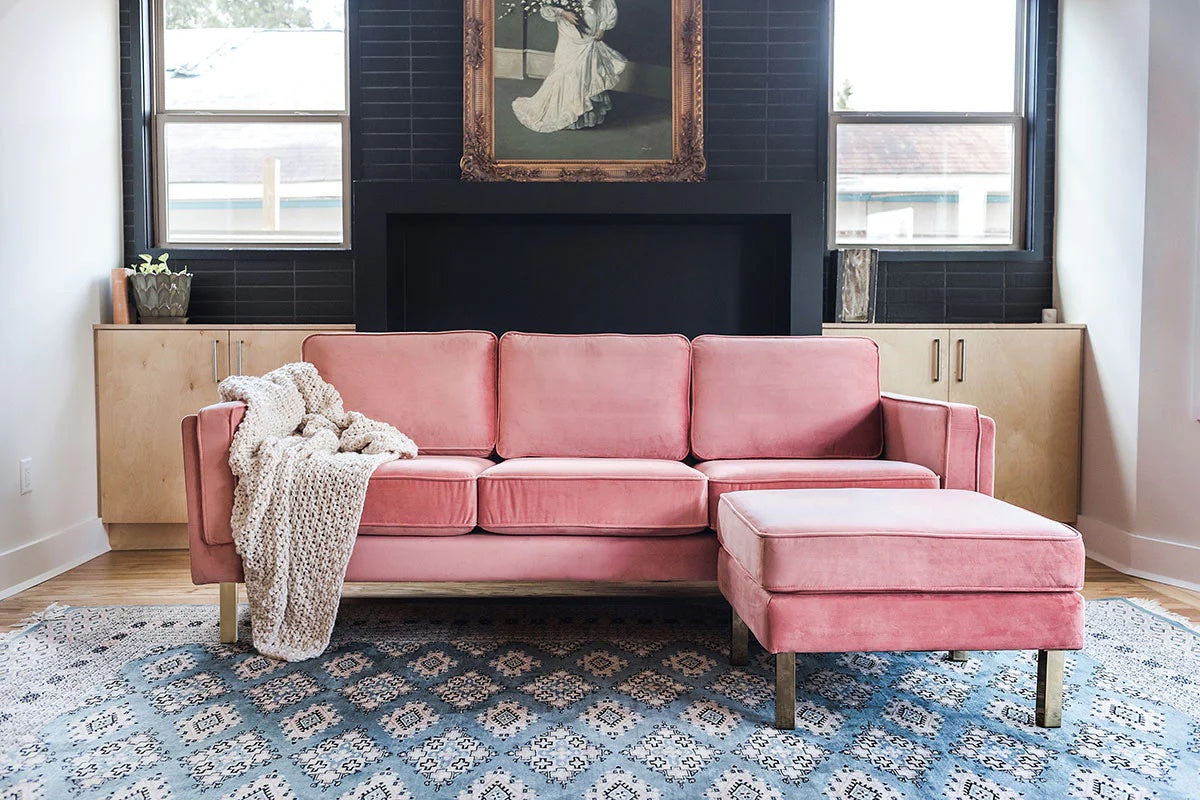 A living room with a pink couch and a matching ottoman on a patterned blue rug. The couch has a beige knit blanket draped over one arm. In the background, there is a fireplace flanked by light wood cabinets and two windows. A framed painting hangs above the fireplace.