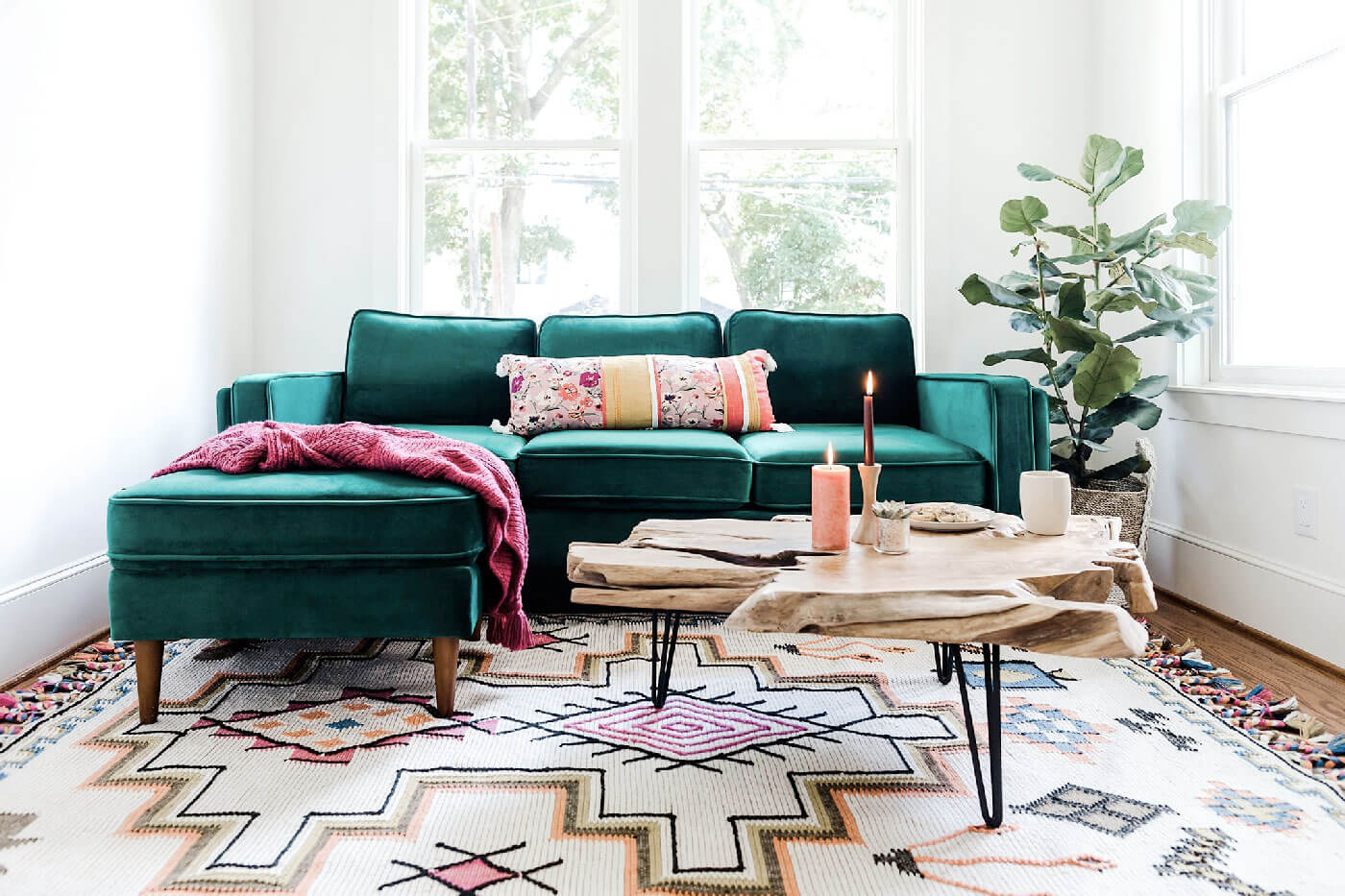 A brightly lit living room features a green sectional sofa adorned with a colorful pillow and a pink throw blanket. In front of the sofa is a wooden coffee table with candles and decorative items. A patterned rug covers the floor, and a large potted plant is seen in the corner.