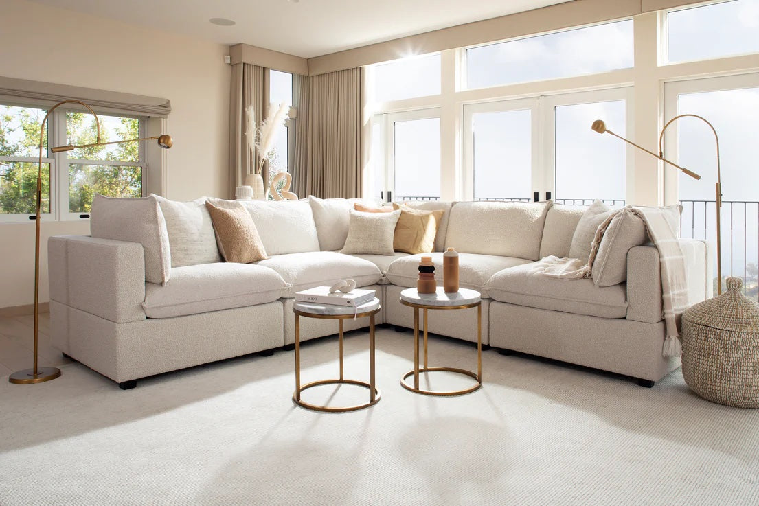 Bright living room with a large beige sectional sofa adorned with neutral-colored cushions. Two round gold coffee tables with decor sit on a light-colored rug. Large windows with curtains allow sunlight to fill the space, and modern floor lamps enhance the ambiance.