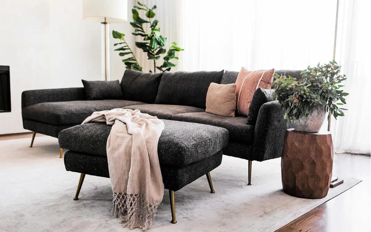 A modern living room featuring a dark gray L-shaped sectional sofa with a matching ottoman. The sofa is adorned with various throw pillows, and a beige blanket is draped over the ottoman. Nearby, there is a large plant and a wooden side table with foliage on it.