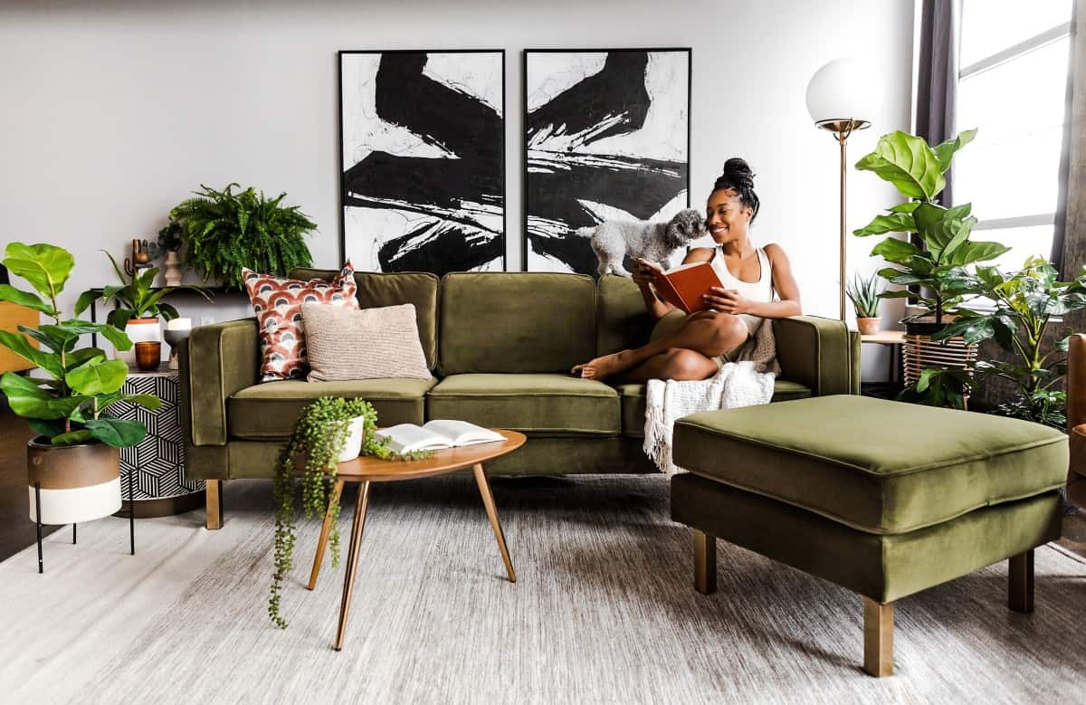 A woman sits on a green sofa reading a book with a cat nearby in a modern living room. The room features various green plants, abstract black and white wall art, a wooden coffee table, and a large ottoman. The space is brightened by a floor lamp and natural light.