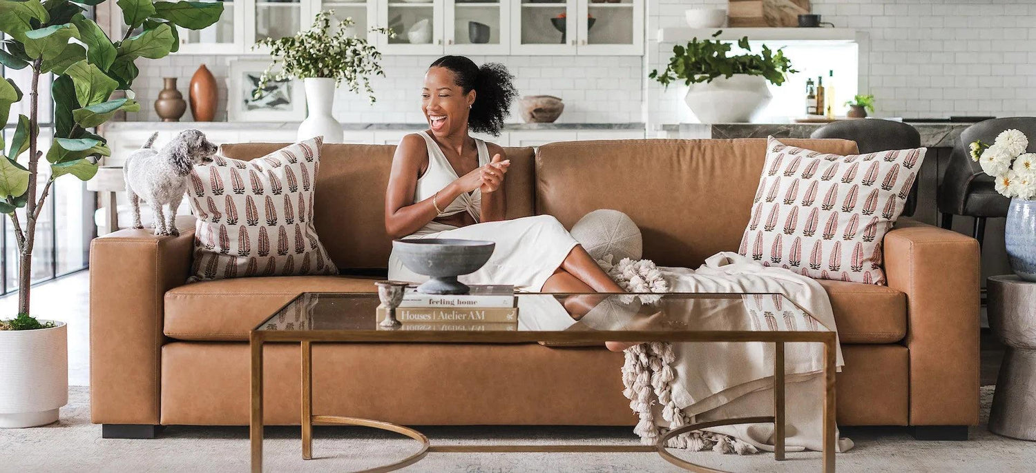 A woman in a white dress sits on a tan leather sofa and laughs, looking at a small dog standing beside her. The living room is decorated with plants, patterned throw pillows, and a glass coffee table with books and decor items. A kitchen is visible in the background.