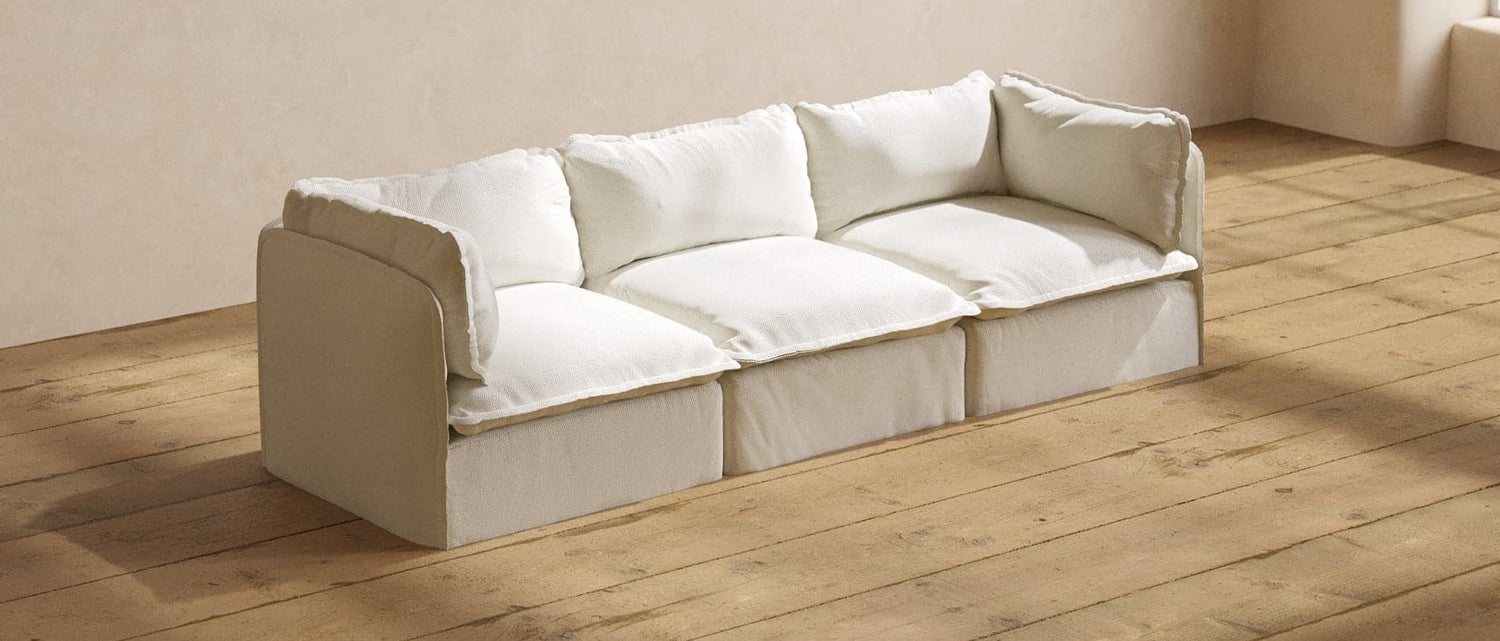 A beige, three-seater sofa with plush white cushions is placed against a neutral beige wall on a wooden floor. The room is well-lit, casting soft shadows on the floor and highlighting the sofa's comfortable and cozy appearance.