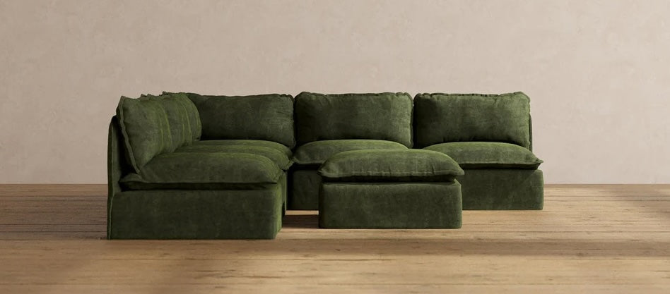 A green sectional sofa with plush cushions is arranged in an L-shape. It includes a matching ottoman, and is set against a plain beige wall on a wooden floor. The furniture has a soft, comfortable appearance.