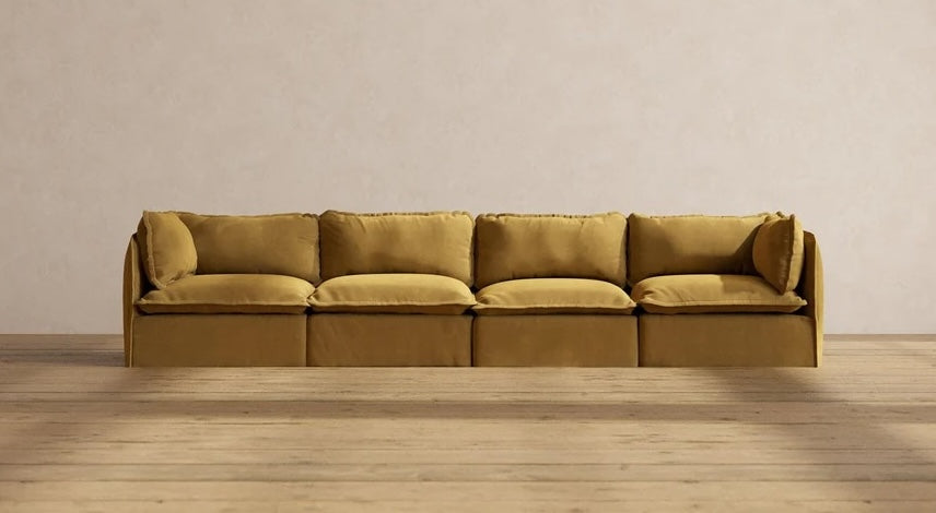 A mustard yellow, modern sectional sofa with four cushioned seats and matching backrest pillows is centered against a plain, light beige wall. The sofa sits on a smooth wooden floor, enhancing the minimalist aesthetic of the room.