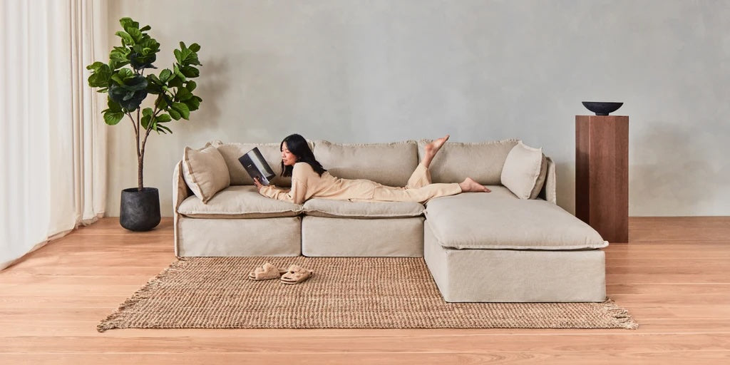 A woman in comfortable clothing lounges on a large beige sectional sofa, reading a book. A jute rug, a pair of slippers, a tall indoor plant, and a wooden pedestal with a bowl on it complete the minimalist living room setting.