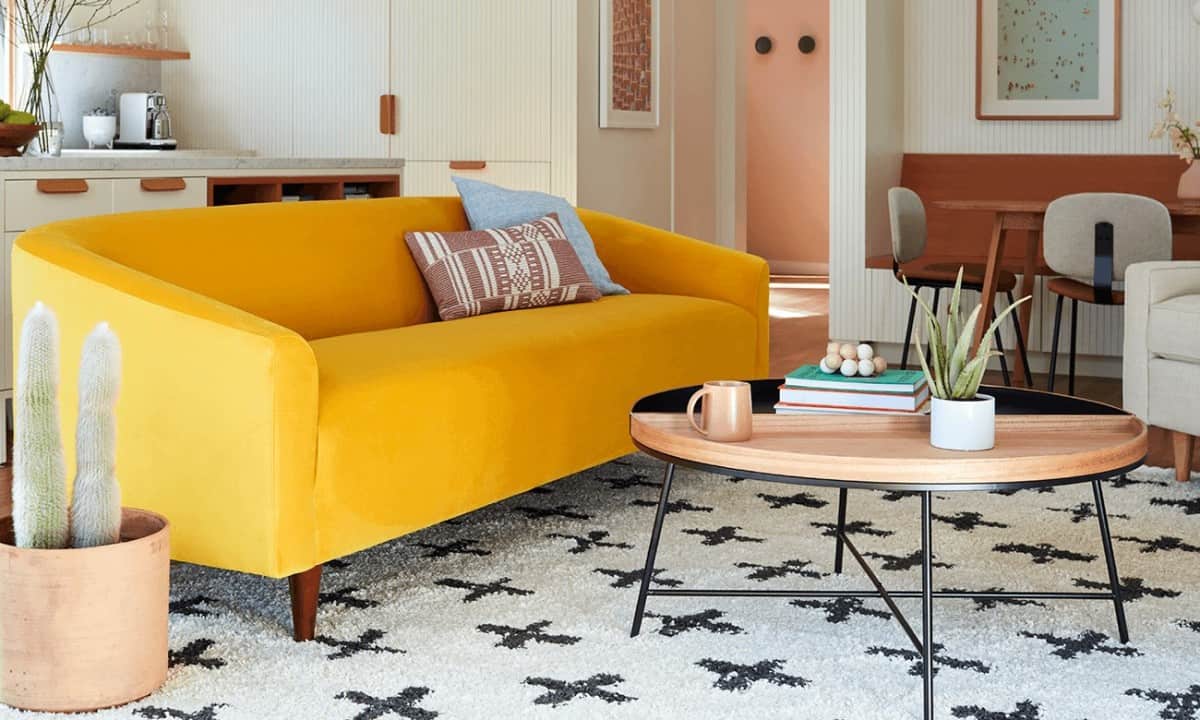 A living room features a mustard yellow sofa adorned with decorative pillows. In front of the sofa is a round wooden coffee table with a black rim, holding books and a potted plant. The light-colored rug has a star pattern, and a tall cactus sits nearby.