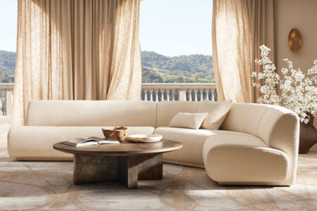 A luxurious living room features a beige sectional sofa and a round wooden coffee table with books and a bowl on it. Large windows with light curtains reveal a scenic view of green hills. A vase of white flowers stands on the right, enhancing the elegant decor.