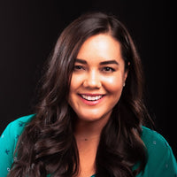 Arlyn Hernandez, writer and editor. A woman with long, dark, wavy hair smiles while looking at the camera. She is wearing a teal top with a pattern of small white and black shapes. The background is black, highlighting her face and outfit.