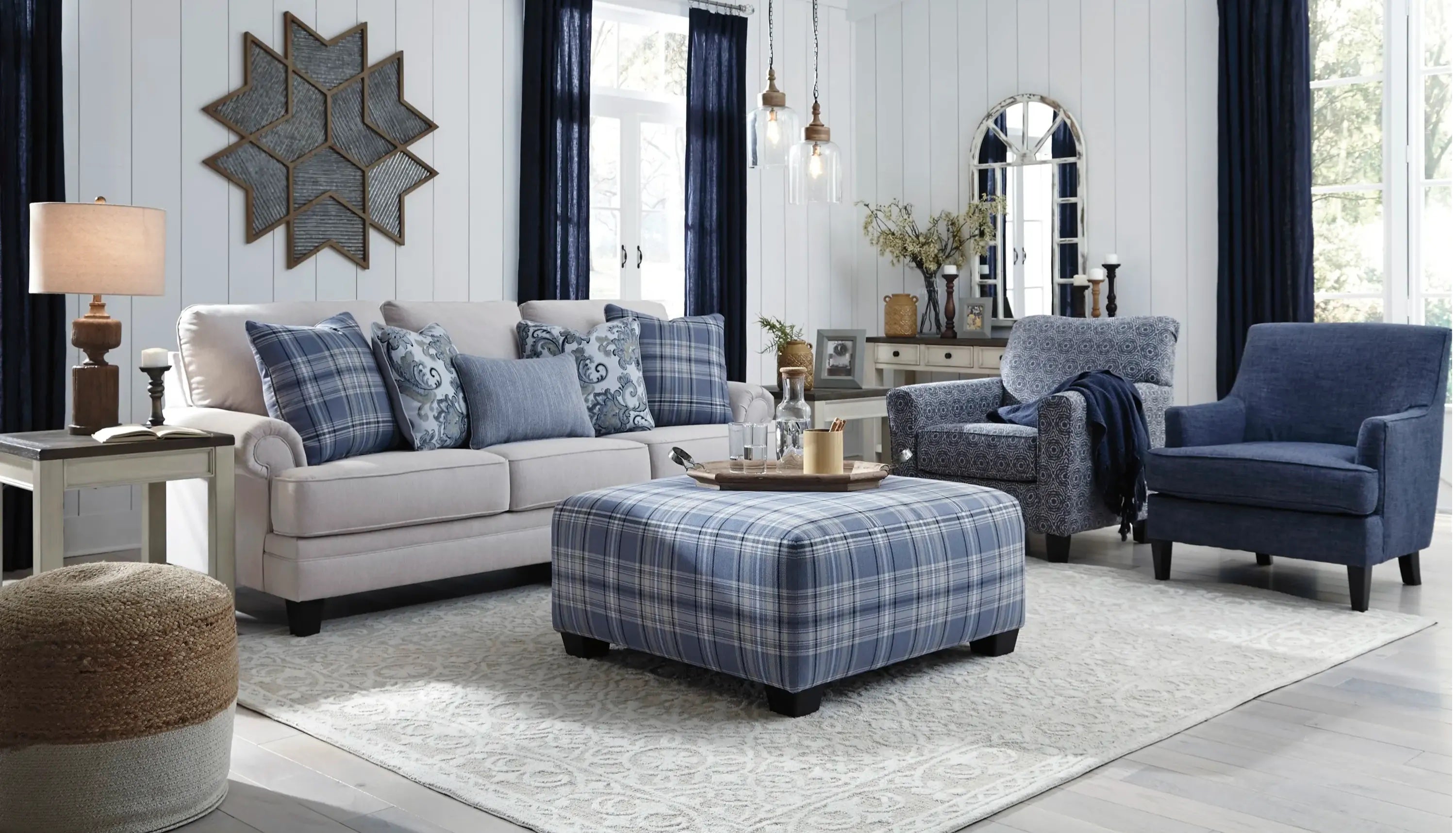 A stylish living room with a light beige sofa adorned with blue plaid and patterned pillows, a large plaid ottoman, a blue upholstered armchair, and a wooden coffee table. The room features dark blue curtains, wall art, a large mirror, and decorative lighting.