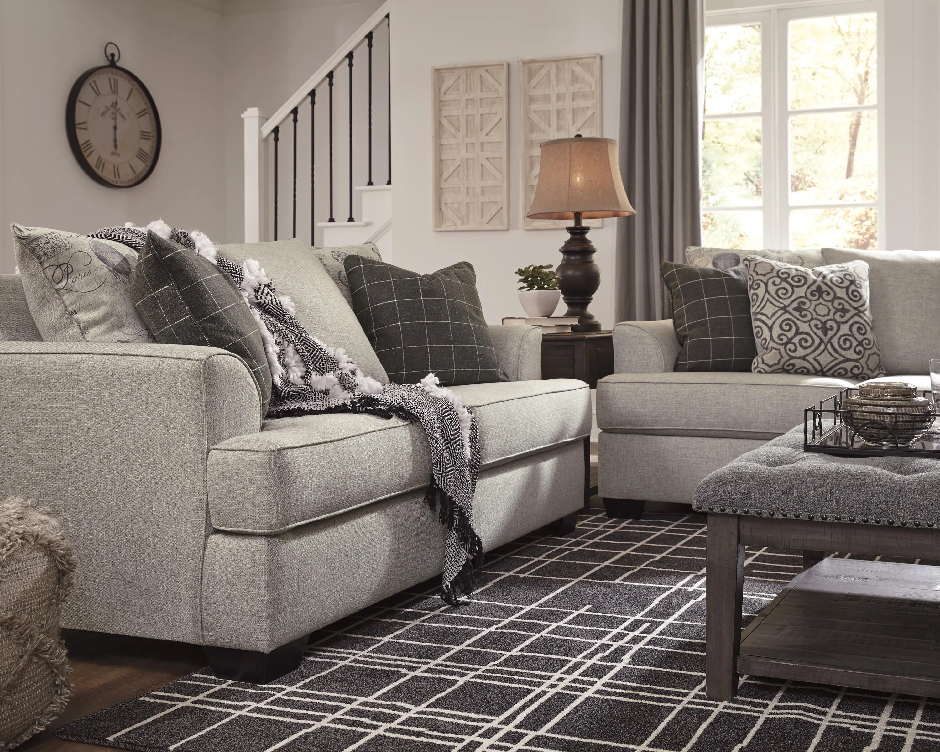 A cozy living room with two gray sofas adorned with patterned pillows and a monochrome throw blanket. A coffee table with a decorative tray sits on a black-and-white geometric rug. A lamp on a side table, abstract wall art, and a large clock complete the decor.