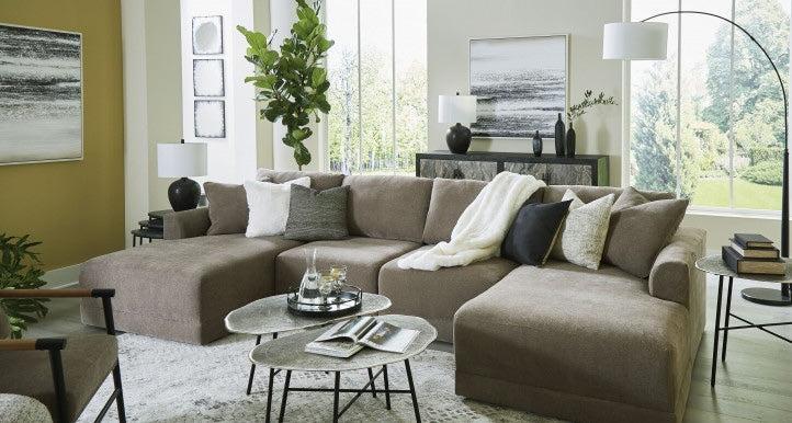 A modern living room featuring a large beige sectional sofa with assorted throw pillows and a white blanket. A set of round, nested coffee tables holds books and decorative items. The room is adorned with large windows, potted plants, and framed artwork.