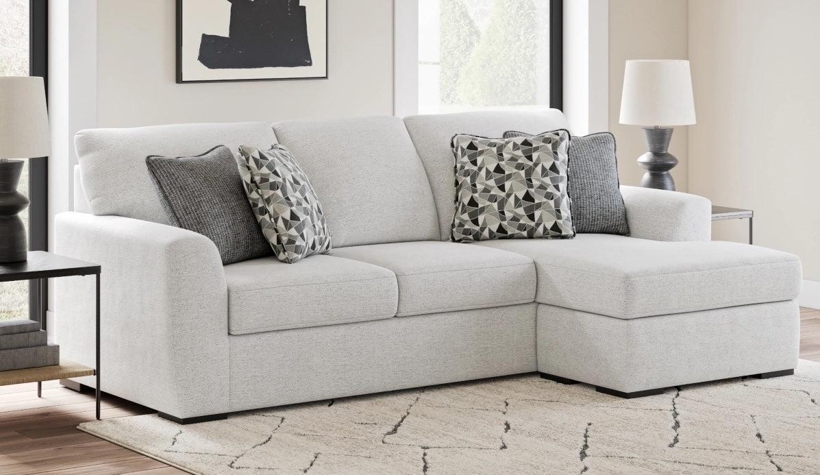 A modern living room featuring a light gray sectional couch. The couch includes gray geometric-patterned and solid gray cushions. It is flanked by two dark wood side tables, each with a white lamp. A light-colored rug lies on the wooden floor.