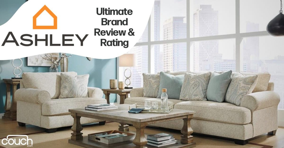 A modern living room setting featuring beige couches with blue and beige cushions, a wooden coffee table with a glass centerpiece, and large windows displaying a cityscape. Text reads "Ashley Ultimate Brand Review & Rating" with the Ashley logo and "couch.com" watermark.