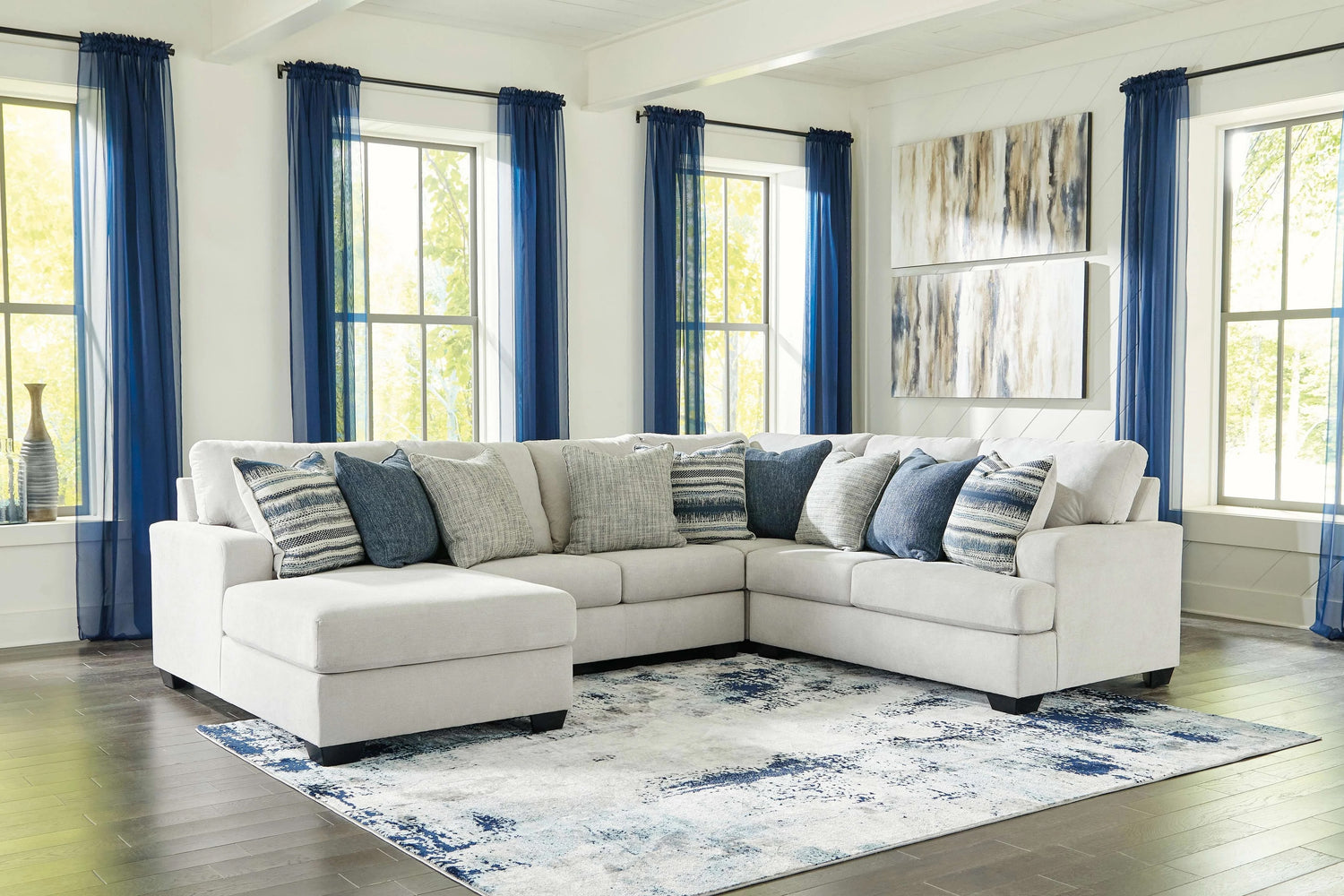 A spacious living room features a large, L-shaped beige sectional sofa adorned with blue and gray throw pillows. The room has large windows with blue curtains, abstract paintings on the wall, and a blue-and-white area rug on the dark hardwood floor.