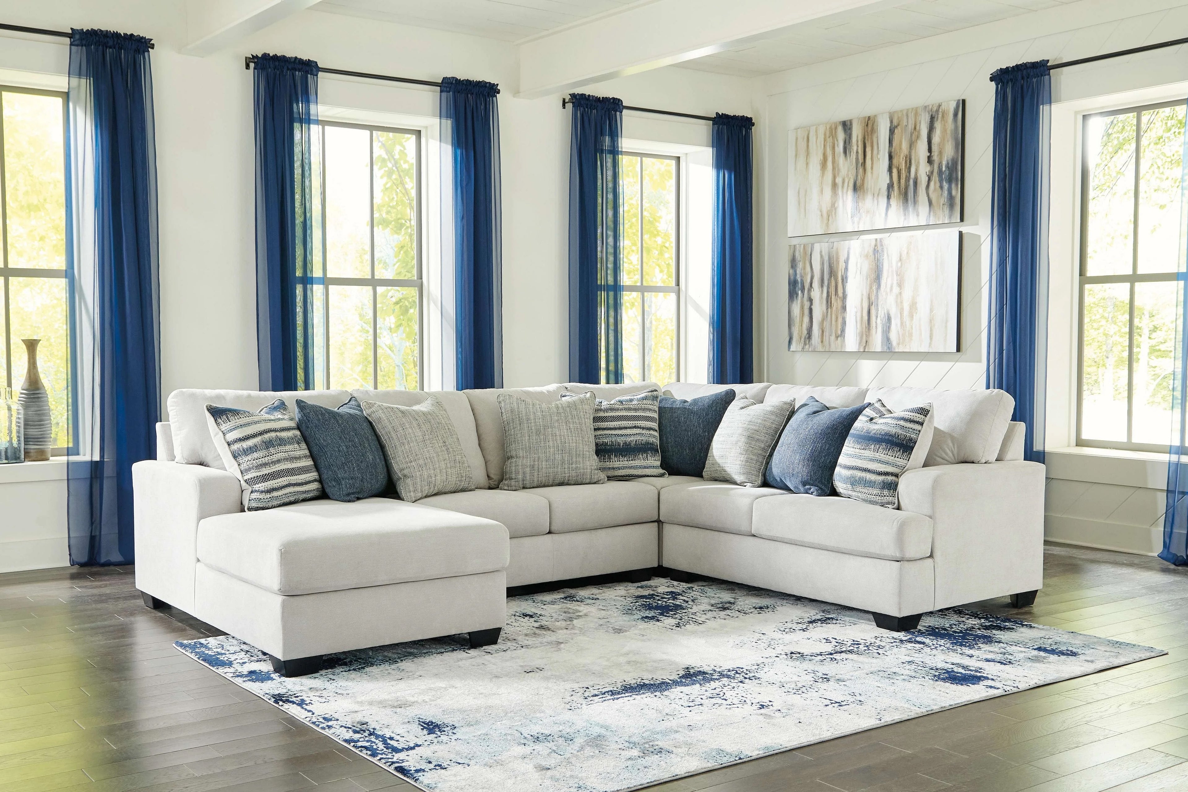 A spacious living room features a large, L-shaped beige sectional sofa adorned with blue and gray throw pillows. The room has large windows with blue curtains, abstract paintings on the wall, and a blue-and-white area rug on the dark hardwood floor.