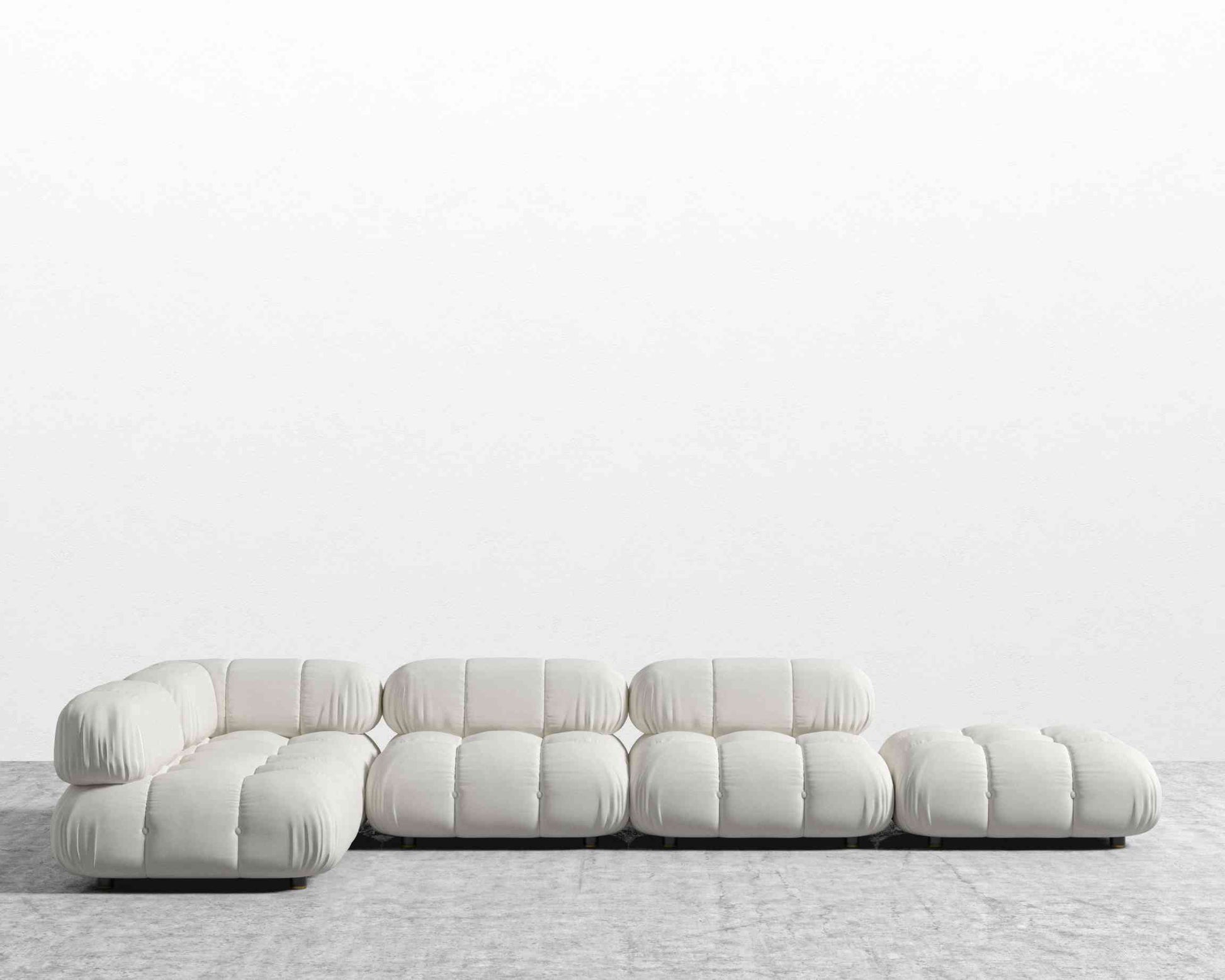 The Belia Modular Sectional, featuring white, tufted cushions, is arranged in an L-shape against a plain white wall on the gray carpeted floor. This sofa design showcases low-profile seating and a minimalist aesthetic.