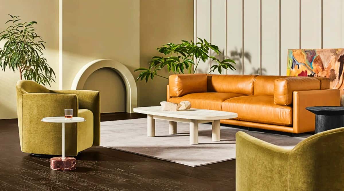 A modern living room featuring a tan leather sofa, two olive green armchairs, and a white coffee table with decorative items. A light gray rug covers the dark wood floor. Plants and a colorful abstract painting add a touch of vibrancy to the space.